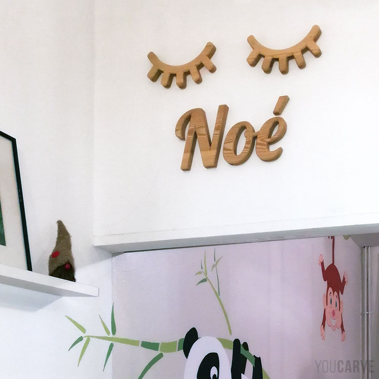 Prénom Noé en lettres découpées en bois (mélèze) avec décoration paupières en bois, fixation mousse double-face sur mur de chambre d'enfant.