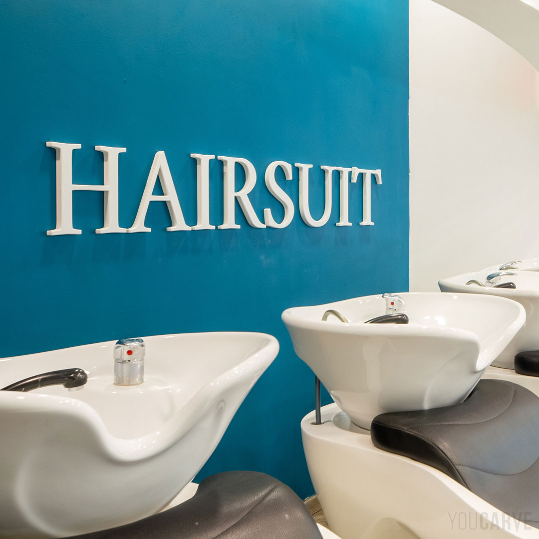 Réalisé par Hairsuit (salon de coiffure à Annecy) : logo mural en relief, découpe sur-mesure en PVC expansé blanc épaisseur 19 mm finition chanfreinée, fixation mousse-double-face avec gabarit de positionnement.